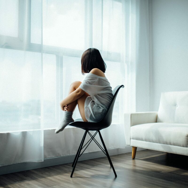 Woman sat on a chair, clutching her knees and staring our of a window feeling sad